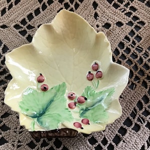 Op de afbeelding: Een gele keramische schotel in de vorm van een blad met groene bladeren en rode bessen die erop zijn geschilderd.