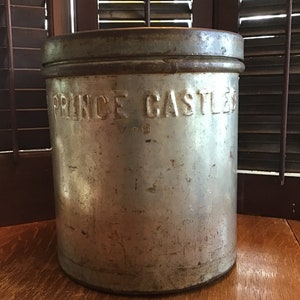 May include: A cylindrical metal container with the embossed text "PRINCE CASTLES". The container has a lid and a slightly rusted appearance. The container is sitting on a wooden surface.