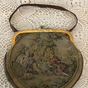 K&ouml;nnte beinhalten: Eine runde, mit Tapisserie bezogene Handtasche mit goldenem Rahmen und einem braunen Lederriemen. Die Tapisserie zeigt eine Szene mit Menschen in einem Garten.