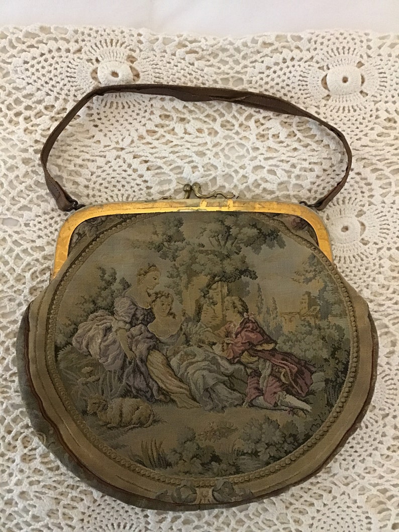 K&ouml;nnte beinhalten: Eine runde, mit Tapisserie bezogene Handtasche mit goldenem Rahmen und einem braunen Lederriemen. Die Tapisserie zeigt eine Szene mit Menschen in einem Garten.