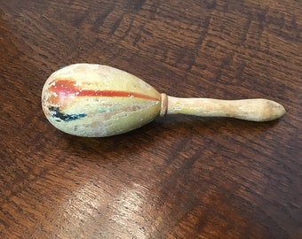 May include: A wooden maraca with a yellow, red, and blue painted design. The maraca has a wooden handle.
