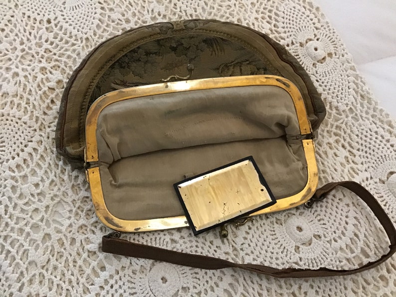 K&ouml;nnte beinhalten: Eine vintage, halbmondf&ouml;rmige Handtasche mit einem goldenen Verschluss und einem braunen Riemen. Die Tasche ist aus einem Stoff mit Blumenmuster und hat einen kleinen, rechteckigen Spiegel im Inneren.