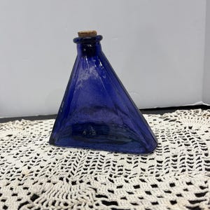 Puede incluir: Una botella triangular de vidrio azul cobalto con tapón de corcho. La botella tiene una forma única y está sobre un tapete de ganchillo color crema. La forma de la botella recuerda a una pirámide.