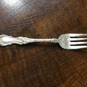 Puede incluir: Un tenedor de plata con un mango adornado y tres dientes. El tenedor está sobre una superficie de madera marrón oscura.