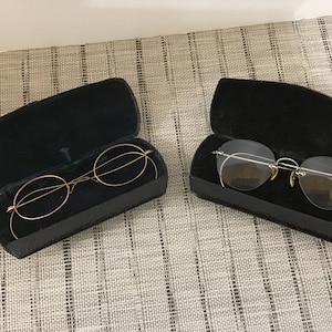 Puede incluir: Dos pares de gafas vintage en estuches de terciopelo negro. Un par tiene gafas redondas con montura dorada. El otro par tiene gafas ovaladas con montura plateada.