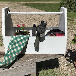 Può includere: Un contenitore per attrezzi in legno bianco con un manico. Il contenitore contiene attrezzi da giardino, una pianta in vaso e un panno a quadri verde e bianco.