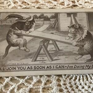May include: A vintage postcard featuring a black and white illustration of two cats on a seesaw. The cat on the left is wearing a hat with a feather. The cat on the right is wearing a cone-shaped hat. The caption reads "I'll join you as soon as I can - I'm doing my best."