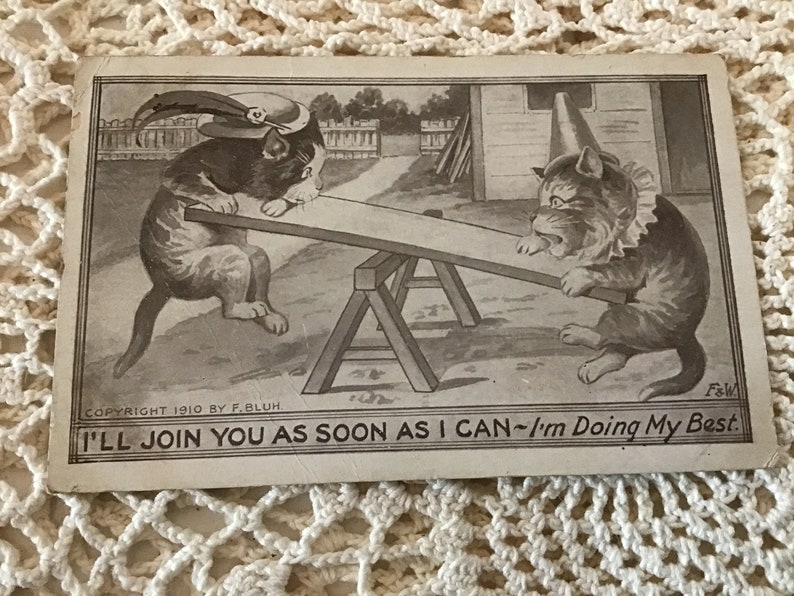 May include: A vintage postcard featuring a black and white illustration of two cats on a seesaw. The cat on the left is wearing a hat with a feather. The cat on the right is wearing a cone-shaped hat. The caption reads "I'll join you as soon as I can - I'm doing my best."