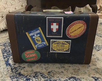 May include: A vintage blue suitcase with brown leather trim and several hotel stickers from Paris, France. The stickers include the names "Hotel Ambassador", "Hotel Sylva", "Hotel de Florence", "Hotel Scribe", and "Dessees Palace - Hotel".