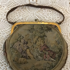 K&ouml;nnte beinhalten: Eine runde, mit Tapisserie bezogene Handtasche mit goldenem Rahmen und einem braunen Lederriemen. Die Tapisserie zeigt eine Szene mit Menschen in einem Garten.