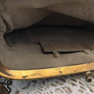 K&ouml;nnte beinhalten: Das Innere einer Vintage-Handtasche mit einem goldenen Metallrahmen und einem braunen Lederriemen. Das Futter ist aus hellem beigefarbenem Stoff mit einer kleinen Tasche.