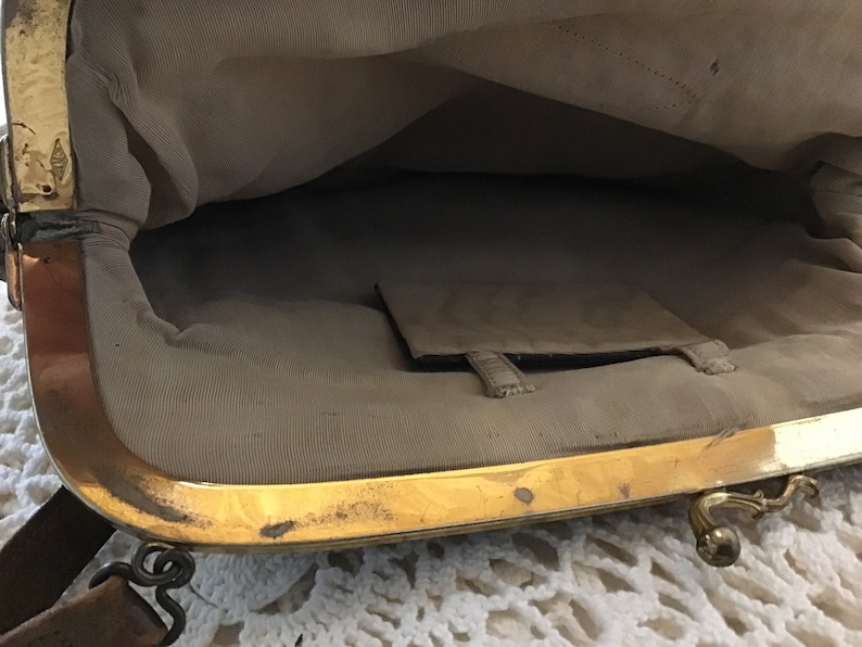 K&ouml;nnte beinhalten: Das Innere einer Vintage-Handtasche mit einem goldenen Metallrahmen und einem braunen Lederriemen. Das Futter ist aus hellem beigefarbenem Stoff mit einer kleinen Tasche.