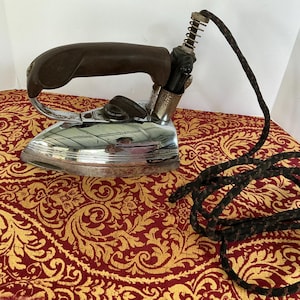 May include: Vintage electric iron with a brown handle and chrome base. The iron has a long, black, braided power cord. The iron is resting on a red and gold patterned fabric.