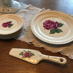 Pode incluir: Um conjunto de pratos de cerâmica branca com borda dourada e design de rosa rosa. Os pratos estão empilhados uns sobre os outros. Há também um servidor de bolo de cerâmica branca com cabo dourado e design de rosa rosa.