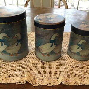 Pode incluir: Três latas redondas de metal com tampas azuis e uma cena de inverno com neve com gansos usando fitas azuis. As latas estão sobre um crochê branco.