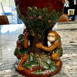 May include: A ceramic vase with a red, brown, and green glaze. The vase is decorated with a tree and two monkeys. The vase has a white top with a crackled glaze.