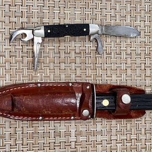 May include: A multi-tool pocket knife with a black handle and silver metal components, including a blade, bottle opener, and awl, positioned above a brown leather sheath containing a second knife with a black handle and brass rivets.