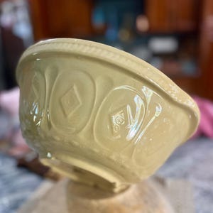 May include: A pale yellow ceramic mixing bowl featuring a raised embossed pattern around the exterior. The design includes repeating diamond shapes and vertical oval motifs, finished with a textured rope-like detail along the thick, rounded rim.