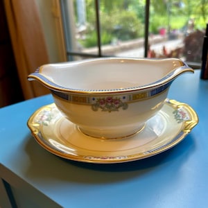 May include: Cream-colored gravy boat and saucer set. The boat has a blue and gold rim with a floral design. The saucer has a matching design and gold trim. A classic addition to any dining table.