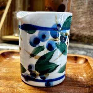 May include: A white ceramic creamer pitcher featuring a hand-painted botanical pattern of dark blue berries and green leaves. The cylindrical vessel has a small pouring spout at the top rim and is decorated with horizontal blue bands near the top and base.