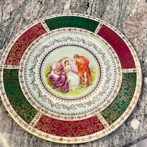 Könnte beinhalten: Ein weißer Porzellanteller mit goldfarbenem Rand und einem zentralen Bild von drei Figuren in einer Gartenlandschaft. Der Teller ist mit einem sich wiederholenden Muster aus grünen, goldenen und roten floralen Designs verziert.