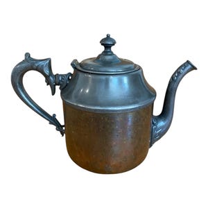 May include: Antique teapot with a copper body and a silver-colored lid, handle, and spout. The teapot has an ornate handle and spout design. The copper body has a textured appearance. The lid is topped with a small knob.