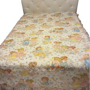 May include: A quilted bedspread featuring a repeating pattern of Cabbage Patch Kids dolls in various outfits and poses, including playing with toys and kites. The fabric has a white background with soft pastel colors and a ruffled bottom edge.