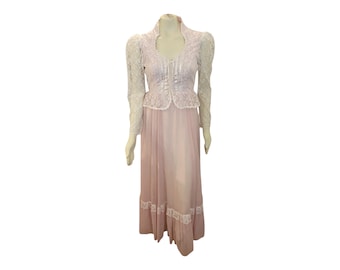 May include: A vintage-style, floor-length dress on a mannequin. The dress features a light pink skirt with a white lace trim at the hem. The bodice is a matching light pink with a white lace overlay and long, white lace sleeves.