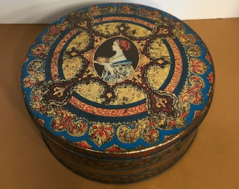 May include: Round vintage tin with a decorative design in blue, gold, and red. The lid features a central portrait of a woman and ornate floral patterns. The tin has a layered side.