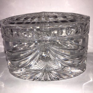 May include: A clear glass oval-shaped trinket box with a lid. The box has a cut glass design with a starburst pattern on the bottom and a decorative band around the top.