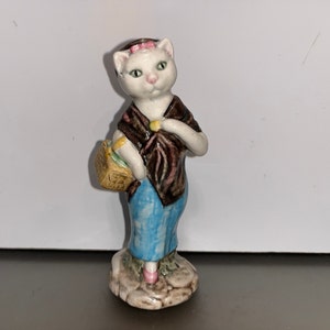 Puede incluir: Una figurita de cerámica blanca de un gato que lleva un vestido marrón y rosa y lleva una cesta. El gato tiene un lazo rosa en el pelo y pantalones azules.