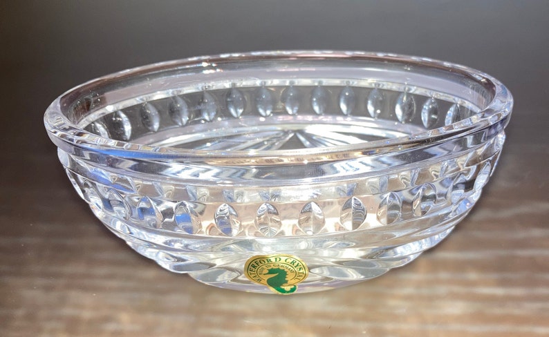 Puede incluir: Un cuenco de vidrio transparente con un patr&oacute;n decorativo de peque&ntilde;os c&iacute;rculos y &oacute;valos. El cuenco tiene una pegatina dorada en la parte inferior con el texto "Waterford Crystal" y un logotipo de caballito de mar.