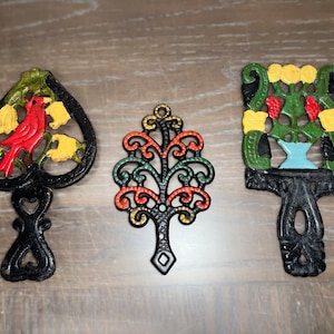 May include: Three vintage cast iron hot pads with colourful designs. The first is a heart shape with a red cardinal bird and yellow flowers. The second is a tree shape with green, red, and gold swirls. The third is a rectangular shape with a blue vase and green and yellow flowers.