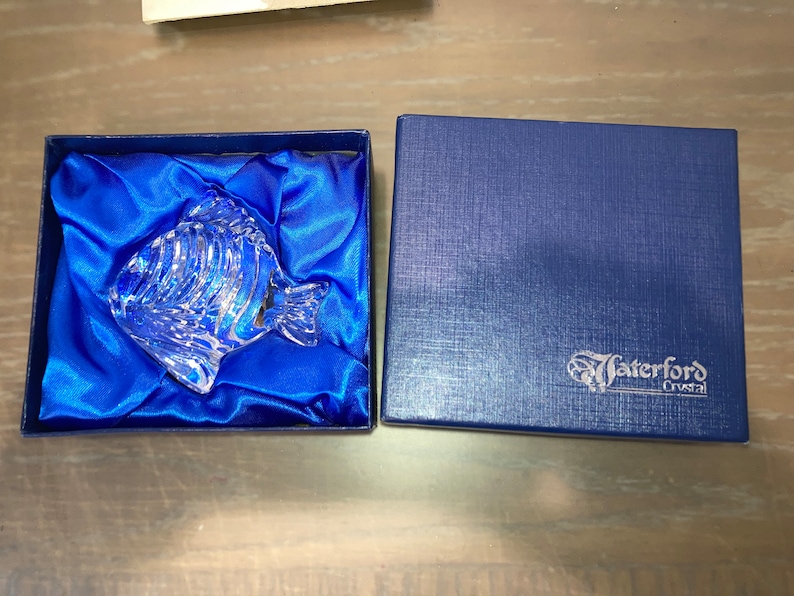 Peut inclure: Une figurine de poisson en verre transparent dans un coffret en velours bleu avec une doublure en satin bleu. Le coffret porte une &eacute;tiquette blanche avec le texte "Waterford Crystal".