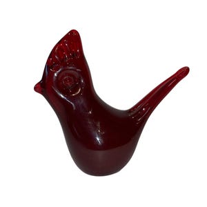 Könnte beinhalten: Glänzende, rubinrote Glasvogelfigur. Der Vogel hat einen detaillierten Kamm, ein rundes Auge und einen langen, spitzen Schwanz. Die Figur ist ein Dekorationsartikel, wahrscheinlich zur Präsentation.