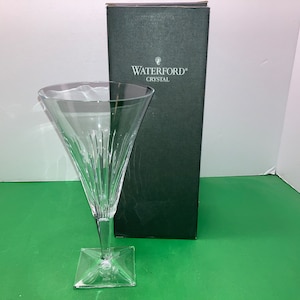 May include: A clear glass Waterford crystal champagne flute with a square base and a delicate, etched design. The flute is sitting on a green surface in front of a black box with the Waterford Crystal logo.