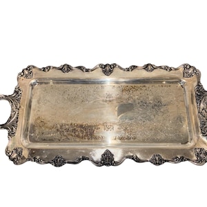 May include: A rectangular silver serving tray with ornate handles and a decorative scalloped edge. The tray has a detailed floral pattern etched into the surface. The handles are curved and feature intricate scrollwork.