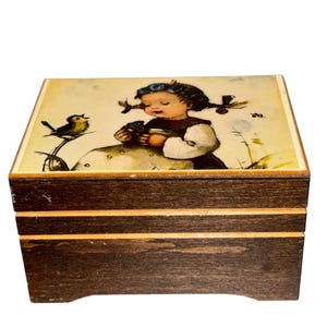 May include: A wooden music box with a hinged lid. The lid features a colorful illustration of a young girl with pigtails, holding a bird, and a small bird perched on a branch. The box has a dark brown finish with a lighter brown accent.