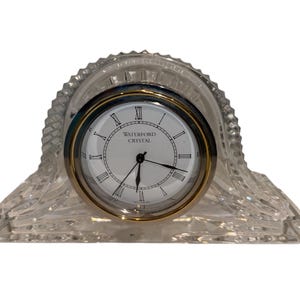 May include: A clear crystal clock with a white clock face and black hands. The clock face has Roman numerals and the words "WATERFORD CRYSTAL". The clock has a gold-colored bezel and a decorative crystal base.