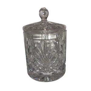 May include: A clear, cut-glass jar with a lid and a round knob on top. The jar has a cylindrical shape with a detailed, symmetrical pattern. The lid fits snugly, and the glass reflects light, highlighting the intricate design. This decorative jar is suitable for storing small items.