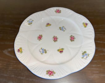 May include: A white ceramic plate with a blue rim and a floral design. The plate has a scalloped edge and is decorated with small pink, yellow, and blue flowers.