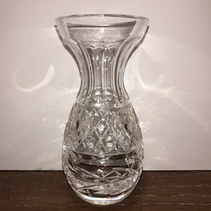 Puede incluir: Un jarrón de cristal tallado transparente, con una parte superior acampanada y una sección inferior con diseño de diamantes. El jarrón tiene una base redondeada y un cuello liso y cilíndrico. El cristal refleja la luz. Probablemente para flores.