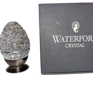 K&ouml;nnte beinhalten: Ein klarer, kristallener, eif&ouml;rmiger Schmuck mit silbernem Sockel. Der Kristall ist facettiert und reflektiert das Licht. Eine graue Schachtel mit den Worten "WATERFORD CRYSTAL" befindet sich neben dem Ornament. Die Schachtel hat ein Seepferdchen-Logo.