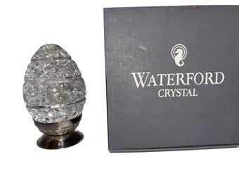 May include: A clear crystal egg-shaped ornament with a silver base. The crystal is faceted, reflecting light. A gray box with the words "WATERFORD CRYSTAL" is next to the ornament. The box has a seahorse logo.
