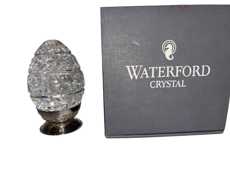 K&ouml;nnte beinhalten: Ein klarer, kristallener, eif&ouml;rmiger Schmuck mit silbernem Sockel. Der Kristall ist facettiert und reflektiert das Licht. Eine graue Schachtel mit den Worten "WATERFORD CRYSTAL" befindet sich neben dem Ornament. Die Schachtel hat ein Seepferdchen-Logo.