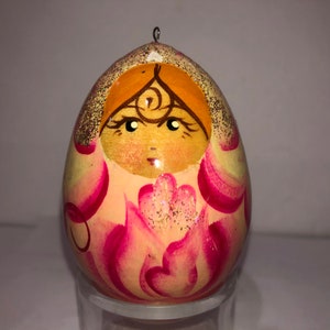 以下が含まれることがあります： ピンクと黄色の花柄が手描きされた木製卵のオーナメント。卵には、王冠をかぶった顔が描かれており、背景は黄色です。