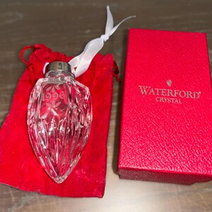 May include: A Waterford Crystal ornament in the shape of a heart with the year 1996 etched on it. The ornament is in a red velvet bag with a white ribbon.