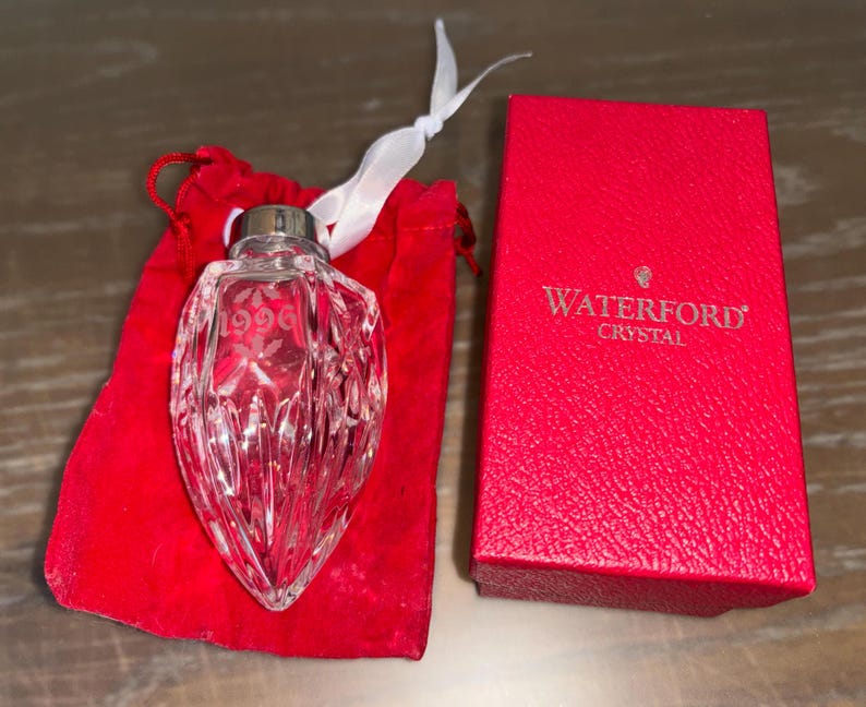 May include: A Waterford Crystal ornament in the shape of a heart with the year 1996 etched on it. The ornament is in a red velvet bag with a white ribbon.