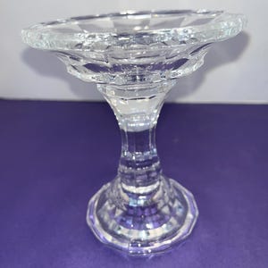 May include: A clear glass candle holder with a wide, scalloped base and a tall, slender stem. The holder has a faceted design.