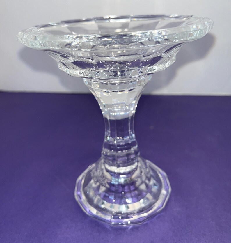 May include: A clear glass candle holder with a wide, scalloped base and a tall, slender stem. The holder has a faceted design.